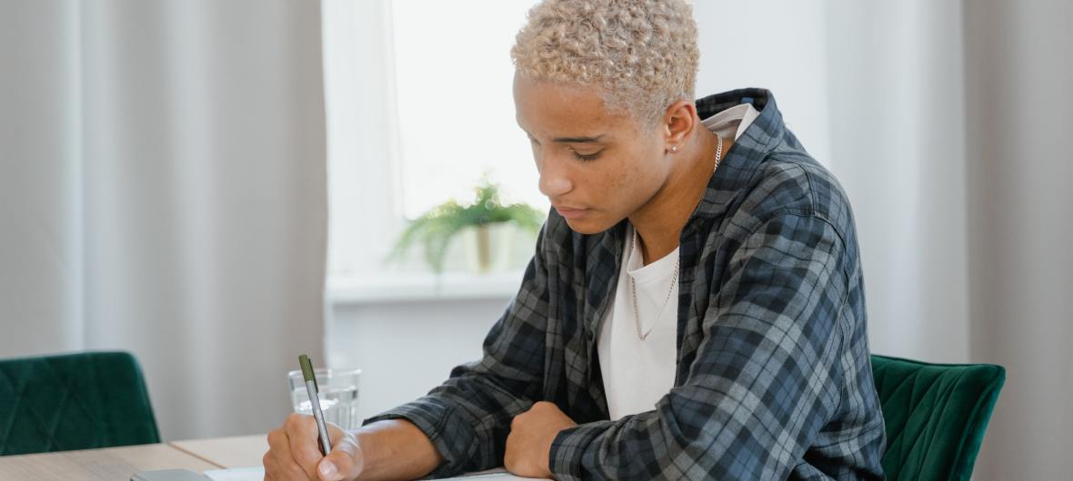 mixed race gender neutral person writing with pen on paper with white background, example of lake superior writers member working on book